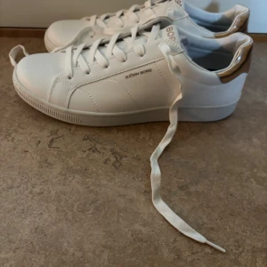 Vita sneakers från Björn Borg - Snygga vita sneakers från Björn Borg med gyllene detaljer på hälen. Skorna har klassisk snörning och en stilren design som passar perfekt till vardags. Tillverkade i syntetmaterial för en modern look.