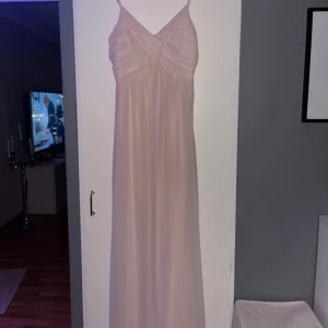 Ljusrosa långklänning från Vila - Elegant ljusrosa långklänning från Vila med smala axelband och v-ringad design. Klänningen har ett fint draperat mönster över bysten och faller vackert ner till golvet. Perfekt för speciella tillfällen.