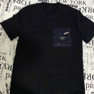 Svart t-shirt med ficka från Prada - Snygg svart t-shirt från Prada med en cool ficka på bröstet som har en dragkedja och logotyp. Perfekt för en stilren look. Kortärmad och i ett mjukt material.och har aldrig använt den pågrund av att den är för stor för mig.