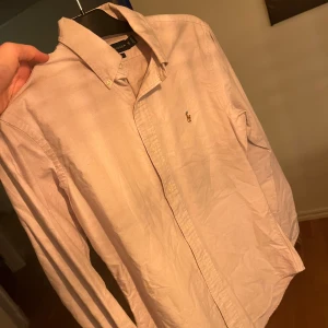 Ljuslila skjorta från Ralph Lauren - Snygg ljuslila skjorta från Ralph Lauren med klassisk krage och broderad logga på bröstet. Skjortan har långa ärmar och knappar framtill. Perfekt för en stilren look.