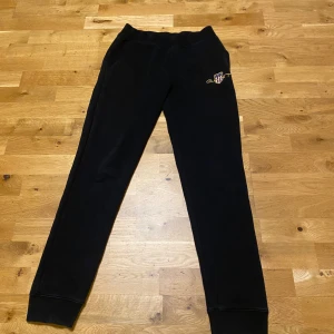 Svarta mjukisbyxor från GANT - Säljer ett par svarta mjukisbyxor från GANT. Byxorna har en enkel design med ribbade muddar vid bensluten och en diskret logga på sidan. Perfekta för en avslappnad stil. Byxorna är i storlek 170cm och i riktigt bra skick, hör av dig via intresse!