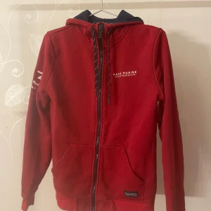 Röd huvtröja från Race Marine - Säljer en snygg röd huvtröja från Race Marine med dragkedja och tryck på ärmen. Tröjan har en justerbar huva med snören och praktiska fickor framtill. Perfekt för en avslappnad stil.