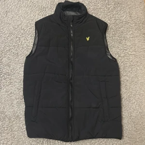 Svart dunväst från Lyle & Scott - Säljer en stilren svart dunväst från Lyle & Scott. Västen har en dragkedja framtill och ett litet broderat emblem på bröstet. Perfekt för kyligare dagar och enkel att matcha med andra plagg.