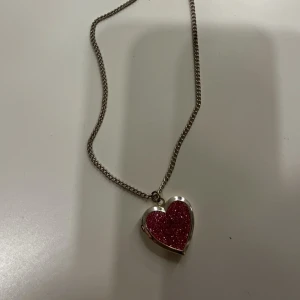 Halsband med hjärtberlock - Charmigt halsband med en hjärtformad berlock som kan öppnas. Berlocken är glittrig i rött och har en silverfärgad kedja. Perfekt för att bära nära hjärtat.