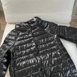 Svart glansig dunjacka - Snygg svart glansig dunjacka med knappar framtill. Jackan har en quiltad design och långa ärmar, perfekt för kyligare dagar. Den har en stilren look som passar till många olika outfits. Herno liknande!