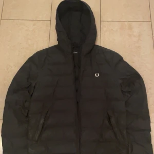 Svart pufferjacka från Fred Perry - Säljer en stilren svart pufferjacka från Fred Perry. Jackan har en huva och dragkedja framtill, perfekt för kyliga dagar. Den ikoniska loggan syns på bröstet. Passar bra för både vardag och mer avslappnade tillfällen. Storlek XS