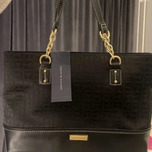 Svart shopperväska från Tommy Hilfiger - Snygg svart shopperväska från Tommy Hilfiger med kedjedetaljer på handtagen och ett subtilt mönster. Väskan har en elegant design med en logotypplatta i metall framtill. Perfekt för att bära med sig alla nödvändigheter med stil. Köpt för lite mer än 1000kr