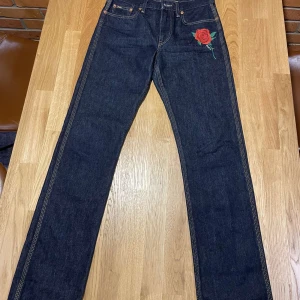 Jeans med rosbroderi från Polo Ralph Lauren - Snygga jeans från Polo Ralph Lauren i mörkblå denim med ett coolt rosbroderi på framsidan. Klassisk femficksdesign med knapp och dragkedja. Perfekta för en stilren look med en touch av färg.