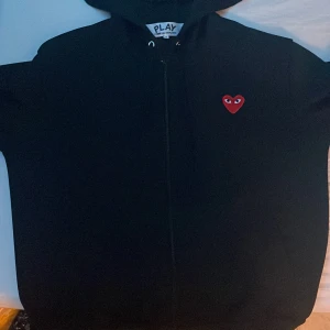 CDG passar S/M - Säljer en stilren svart hoodie från Comme des Garçons med det ikoniska röda hjärtat med ögon på bröstet. Hoodien har dragkedja och en bekväm passform. Perfekt för en casual look.
