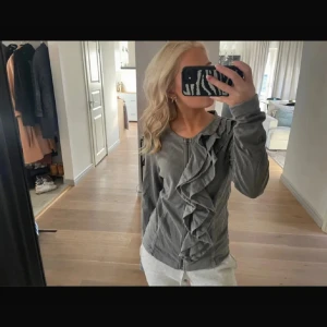 Grå tröja med volang från Esprit - Säljer en stilren grå tröja från Esprit med en snygg volangdetalj framtill. Tröjan har långa ärmar och en dragkedja vid halsen. Perfekt för en chic och avslappnad look.