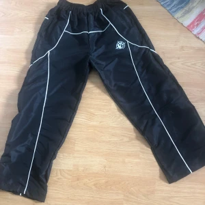 Svarta trackpants från vicinity - Svarta täckbyxor med vita kontrastsömmar och elastisk midja. Byxorna har en loose passform och är perfekta för kalla dagar. Snyggt broderad logga framtill och sportig vibe. Materialet är syntet och de är fodrade för extra värme.