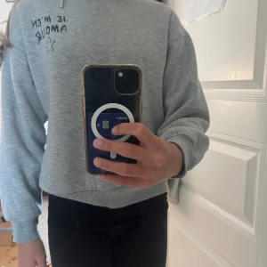 Grå hoodie från Gina Tricot med tryck - Grå hoodie från Gina Tricot med svart text 'JE M'EN AMOUR' och en stjärna tryckt på både fram- och baksidan. Tröjan har huva, långa ärmar och ribbade muddar. Mjuk insida och avslappnad passform, perfekt för chill dagar.