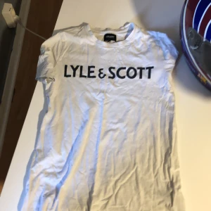 Vit Lyle & Scott t-shirt junior - Vit t-shirt från Lyle & Scott Junior med svart logotyptryck över bröstet. Klassisk rund hals och korta ärmar. Tillverkad i mjuk bomull, perfekt för en chill och enkel stil.