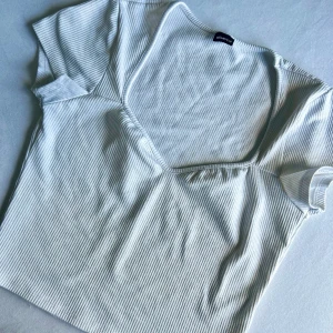 Vit ribbad topp från Gina Tricot - Kortärmad topp i vit ribbad bomull från Gina Tricot. Toppen har en v-ringning och smalrandigt mönster som ger en fräsch och stilren look. Perfekt till jeans eller kjol för en enkel men snygg outfit.