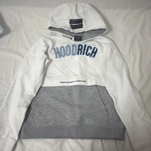 Vit och grå hoodie från Hoodrich 13-15 år - Snygg hoodie från Hoodrich i vitt och grått med blå HOODRICH-tryck på bröstet och texten 'FROM NOTHING TO SOMETHING' vid magen. Stor känguruficka och huva med grå insida. Perfekt för en chill streetwear-look.