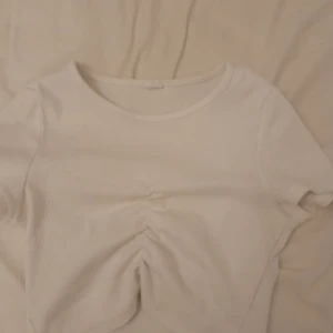 Vit ribbad croppad topp - Säljer en vit ribbad croppad topp med korta ärmar och snygg rynkdetalj framtill. Perfekt för dig som gillar en enkel men trendig stil. Toppens material är mjukt och stretchigt för en bekväm passform.