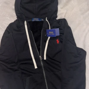Svart hoodie från Polo Ralph Lauren - Svart hoodie från Polo Ralph Lauren med vit snörning och dragkedja framtill. Klassisk röd logga broderad på bröstet. Mjuk bomullskänsla och huva. Perfekt för chill dagar eller när du vill ha en clean streetstyle. Säljer den eftersom den är för liten för mig. Aldrig använt.