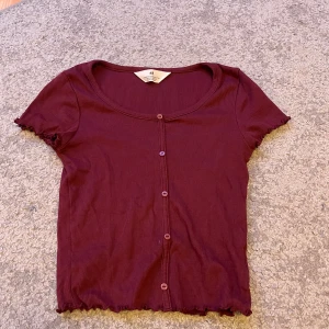 Vinröd ribbad topp med knappar  - Vinröd ribbad topp från H&M med korta ärmar och söt volangkant vid ärmslut och nederkant. Toppen har rund halsringning och knappar framtill, vilket ger en chill och trendig vibe. Perfekt att matcha med jeans eller kjol.
