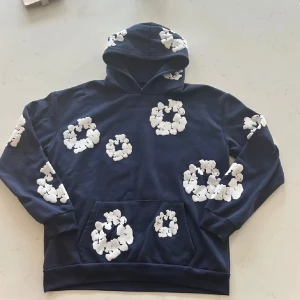 Denim tears  - Säljer en mörkblå hoodie från Denim Tears med vita blommiga mönster över hela tröjan. Tröjan har huva, magficka och långa ärmar. Perfekt för dig som vill sticka ut med en unik och trendig stil.