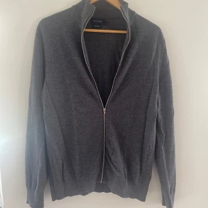 Grå merinoull zip från Riley - Mörkgrå helzip från Riley i mjuk merinoull. Klassisk modell med hel dragkedja framtill och hög krage. Perfekt för lager-på-lager och passar till det mesta. Långärmad och stilren design som funkar året runt.