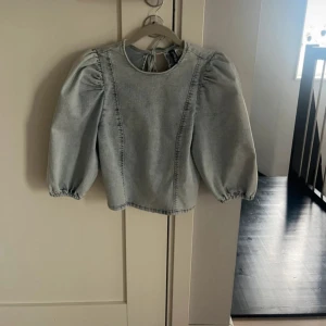 Ljusblå jeansblus med puffärm och knyt - Trendig ljusblå blus i jeanstyg med puffiga trekvartsärmar och rund hals. Baksidan har öppen rygg med två snygga knytband. Perfekt för dig som gillar unika detaljer och en cool, avslappnad look.