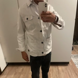 Vit overshirt med stora fickor - Snygg vit overshirt med fyra stora fickor och svarta knappar framtill. Jackan har klassisk krage och är långärmad, perfekt för lager-på-lager. Tillverkad i bomull för en skön och avslappnad känsla.