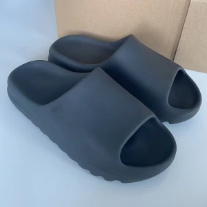 Svarta Yeezy Slides sandaler - Chunky svarta Yeezy Slides från adidas med bred rem och öppen tå. Tillverkade i syntetmaterial med vågformad sula för extra grepp och en clean, minimalistisk look. Perfekta för chill dagar eller när du vill ha en trendig vibe. De har skavanker kolla bilderna.