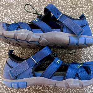 Blå sportiga sandaler med snörning - Snygga blå sandaler med  grå sula och justerbara kardborreband. Elastisk snörning med svarta och gula detaljer samt öppna partier för extra ventilation. I princip oanvända så mycket bra skick. 