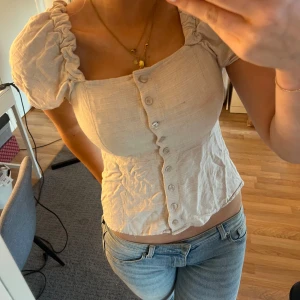 Beige blus med puffärmar och knappar - Supersöt beige blus med puffiga korta ärmar och snygg knäppning framtill. Toppen har en croppad passform och lätt rynkade detaljer vid axlar och midja. Perfekt för dig som gillar romantisk och trendig stil. Tillverkad i ett lätt och luftigt material.