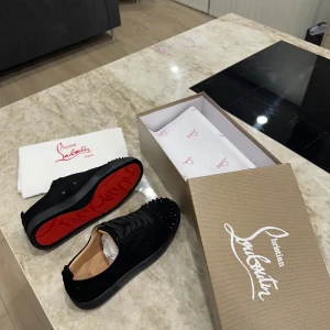 Svarta Louboutin sneakers med nitar - Snygga svarta sneakers från Christian Louboutin med ikonisk röd sula och svarta nitar på tån. Skorna är i mocka och har snörning framtill. Perfekta för dig som vill sticka ut med en lyxig och edgy stil.