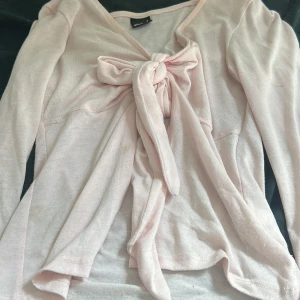 Ljusrosa omlottkofta från Gina Tricot - Supersöt ljusrosa omlottkofta från Gina Tricot med lång ärm och stor knytrosett framtill. Mjuk och tunn i bomullsmix, perfekt för lager-på-lager. Snygg v-ringning och feminin passform som lyfter varje outfit.