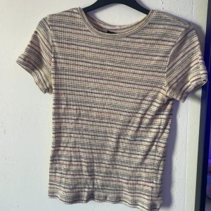 Randig ribbad topp från BikBok - Kortärmad topp från BikBok i ribbat material med smala horisontella ränder i grått, rosa, gult och orange på en ljus bas. Rund halsringning och figurnära passform, perfekt för en chill och färgglad vardagslook.