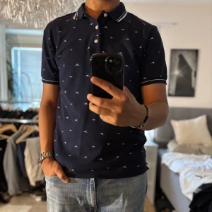 Mörkblå pikétröja Zara Man M - Snygg mörkblå pikétröja från Zara Man i storlek M. Tröjan har små vita broderade fåglar över hela, klassisk krage med vita ränder och tre knappar framtill. Tillverkad i bomull och har korta ärmar med vit rand vid ärmslutet. Perfekt för en clean och stilren look.