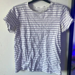 Randig t-shirt från FSBN Sister - Vit och ljuslila randig t-shirt från FSBN Sister. Klassisk rund halsringning och korta ärmar. Tillverkad i mjuk bomull, perfekt för en chill och avslappnad stil. Passar dig som gillar basic men ändå vill ha lite mönster.