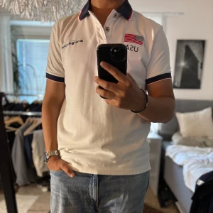 Vit pikétröja Sail Racing USA - Vit pikétröja från Sail Racing med broderad USA-flagga och texten UNITED STATES på ryggen. Mörkblå krage med röd detalj, knappar framtill och broderade loggor. Klassisk passform och korta ärmar, perfekt för en sportig och clean look.