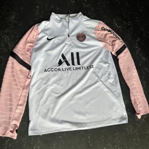 PSG träningströja  - Snygg PSG fotbollströja från Nike med vit kropp och ljusrosa långärmade ärmar. Halv dragkedja framtill, svarta detaljer och klubbmärke på bröstet. Tillverkad i lätt och ventilerande funktionsmaterial, perfekt för träning eller match. M och passar S också , skriv för mer info! 
