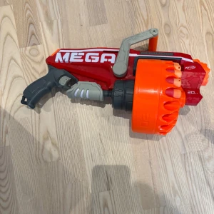 Nerf gun (15 av 20 skott) - Bilderna visar en leksakspistol av typen Nerf Mega Megalodon, inte en bok. Den har en stor orange trumma och plats för flera skumgummipilar. Perfekt för actionfyllda Nerf-strider med vänner!