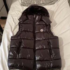 Svart dunväst från Moncler med huva - Säljer en svart, glansig dunväst från Moncler med avtagbar huva och två dragkedjefickor framtill. Västen har klassisk quiltad design och Moncler-logga på bröstet. Perfekt för lager-på-lager och streetstyle.