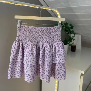 Ljuslila blommig volangkjol Gina Tricot - Supersöt kort volangkjol från Gina Tricot i ljuslila med små blommor. Kjolen har smock i midjan och två volanger nertill. Storlek S