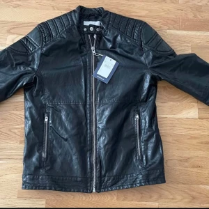 Svart skinnjacka från Springfield L - Svart skinnjacka från Springfield i storlek L. Jackan har coola quiltade detaljer på axlarna, ståkrage med tryckknappar och dragkedja framtill. Två fickor med dragkedja och stilren look som passar till allt. Perfekt för dig som gillar edgy stil.