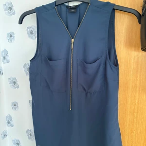 Mörkblå topp i blusmaterial från Lindex i storlek 36 - Jättefin mörkblå topp i blusmaterial från Lindex i storlek 36 med två bröstfickor och en guldig dragkedja framtill. Toppen har en rak passform och är tillverkad i ett luftigt blusmaterial. Köparen står alltid för frakten 