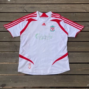 Liverpool bortatröja Adidas vit/röd - Liverpool FC bortatröja från Adidas med klassiskt Carlsberg-tryck på bröstet. Tröjan är vit med röda detaljer och Adidas ränder på axlarna. Broderat klubbmärke och Adidas-logga framtill. Tillverkad i lätt och ventilerande polyester.