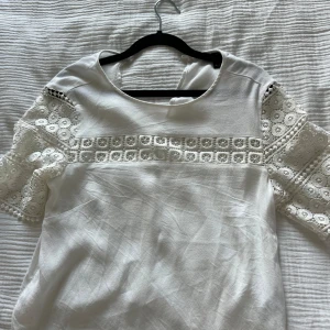 Vit blus med spetsdetaljer - Superfin vit blus med korta ärmar i spets och broderade detaljer över bröstet och ärmarna. Blusen har rund halsringning och ett luftigt, lätt material som känns fräscht och somrigt. Perfekt för dig som gillar romantiska och feminina vibes.