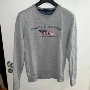 Grå sweatshirt GANT med USA-flagga - Grå sweatshirt från GANT med broderad amerikansk flagga och texten 'GANT 1949' på bröstet. Tröjan har rund hals, långa ärmar. Perfekt för en chill och stilren look.