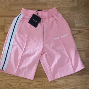 Palm Angels rosa shorts med vit rand - Palm Angels shorts i ljusrosa med vit och svart rand längs sidan. Elastisk midja och fickor på sidorna. Klassisk logga tryckt framtill. Perfekta för sommaren helt nya pris kan diskuteras 