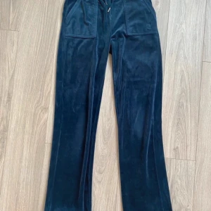 Mörkblå velourbyxor Juicy Couture XS - Mörkblå velourbyxor från Juicy Couture med snörning i midjan och stora fickor fram och bak. Byxorna har en loose passform och raka ben, perfekta för en chill och trendig look. Klassisk Juicy-stil med mjukt material och snygga detaljer.