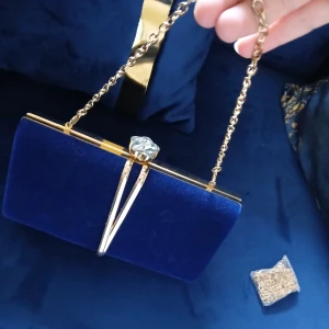 Blå sammet kuvertväska med guldkedja - Säljer en elegant kuvertväska i djupblå sammet med guldfärgade detaljer och kedja💙 Väskan har en stor glittrig stenkristall som lås och insida i svart tyg👛💎Perfekt för att lyfta din outfit med en lyxig touch✨️