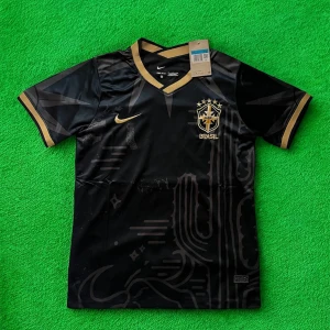 Svart Brasil Nike fotbollströja guld - Snygg svart Brasilien fotbollströja från Nike med guldiga detaljer och broderat CBF-märke på bröstet. Tröjan har V-ringning, korta ärmar och är tillverkad i ett lätt, andningsbart material. Guldiga kanter på ärmar och krage ger en exklusiv känsla.