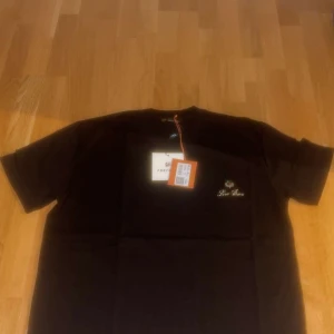 Svart t-shirt från Loro Piana - Svart t-shirt från Loro Piana med broderad logga och emblem på bröstet. Klassisk rund hals och korta ärmar. Skön och stilren modell som passar perfekt till jeans eller shorts. Materialet känns mjukt och exklusivt.