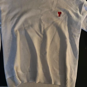 Vit stickad tröja från Ami Paris - Vit stickad tröja från Ami Paris med röd broderad logga (hjärta och A) på bröstet. Tröjan har ribbade muddar och nederkant, klassisk passform och är perfekt för dig som gillar stilren design med en twist.